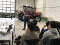 Referent steht vor Zuhörern in Halle, hinter ihm Traktor