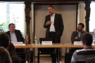 Zwei Männer sitzen an Podium, dazwischen steht Christian Schramm und spricht